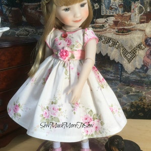 May include: A porcelain doll wearing a white dress with pink floral print and a pink sash. The doll has blonde hair and blue eyes. The doll is wearing pink shoes and is standing on a pink and brown rug. A small black dachshund figurine is sitting on the rug.