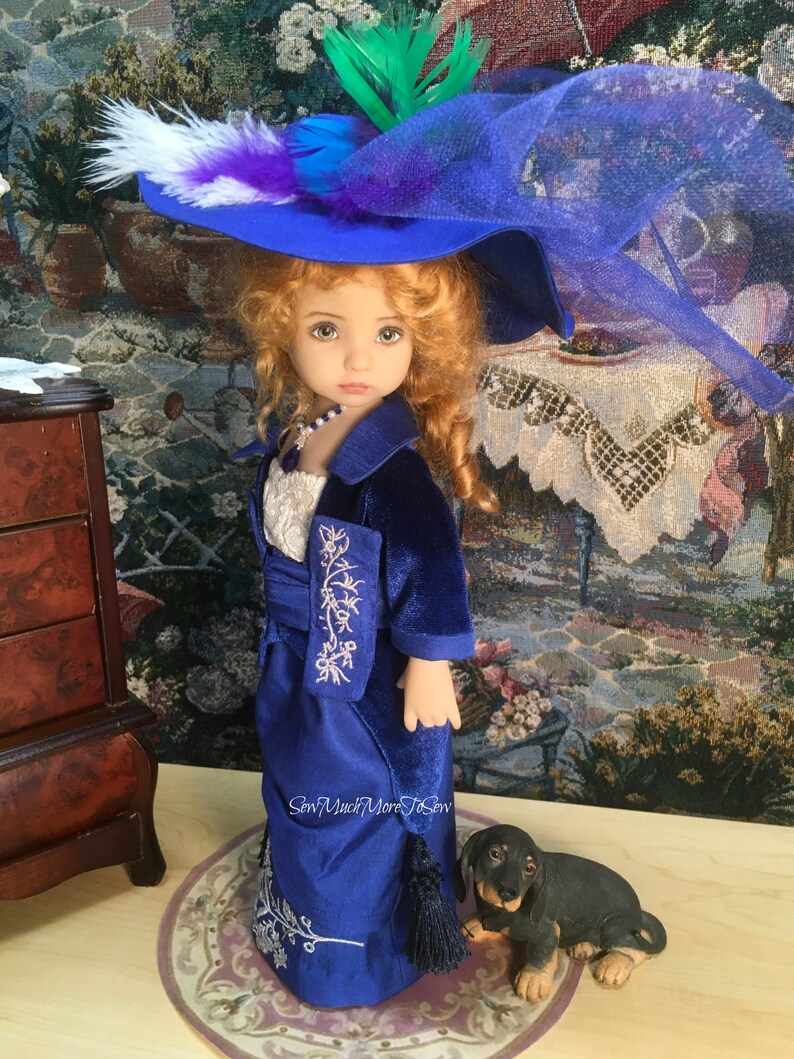 May include: A porcelain doll wearing a blue velvet dress with silver embroidery and a blue hat with a white and purple feather. The doll is standing on a rug with a small black dachshund figurine beside it.