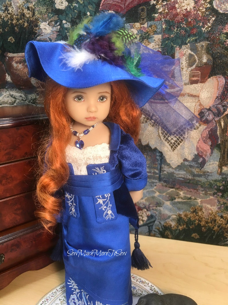 May include: A doll wearing a blue dress with white embroidery and a blue hat with feathers. The doll has long red hair and blue eyes. The doll is wearing a necklace with a blue gem.