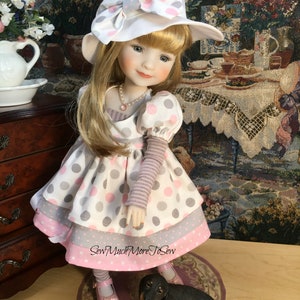 May include: A porcelain doll wearing a white and pink polka dot dress with a matching hat. The doll has blonde hair and blue eyes. The doll is standing on a wooden surface with a small black dog figurine beside her. The text "Sew Much More To Sew" is visible on the doll's dress.