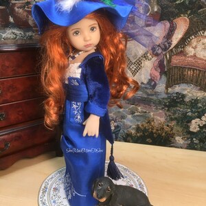 May include: A porcelain doll wearing a blue velvet jacket and a blue satin dress with white embroidery. The doll has long red hair and is wearing a blue hat with feathers. The doll is standing on a round, white, floral patterned surface with a black dachshund figurine.