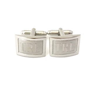 May include: A pair of silver cufflinks with a curved rectangular shape. The cufflinks have the letters "TRL" engraved on them.