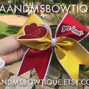 Può includere: Un fiocco da cheerleader rosso e oro con un bordo bianco. Un lato presenta un cappello da cowboy rosso glitterato e l'altro lato dice "yee haw".