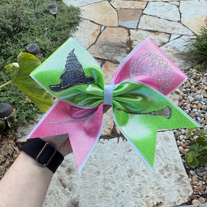 May include: A large, glittery hair bow in pink and green, with witch-themed designs. The bow features a black witch's hat, a broom, and a brain graphic. The bow is held up against a background of greenery and stone.