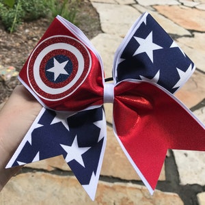 Puede incluir: Lazo de animadora rojo, blanco y azul con un escudo de Capitán América con brillantina en un lado y estrellas en el otro.