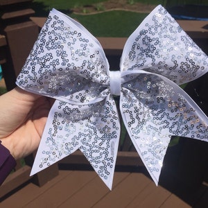 May include: A white cheer bow with silver sequins. The bow is tied in a large, symmetrical knot with two long tails.