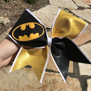 Puede incluir: Lazo de animadora negro y dorado con borde blanco y un logotipo de Batman negro en el centro.