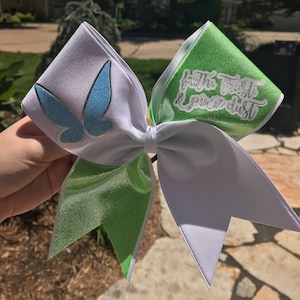 Op de afbeelding: Een witte en groene cheer bow met een blauwe vlinder en de tekst "Faith, Trust & Pixie Dust".