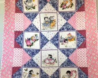 May include: A pink and blue patchwork quilt with a white background. The quilt features a repeating pattern of squares with images of kittens in various poses. The kittens are wearing dresses and are holding objects such as balls of yarn, fish bowls, and butterflies.
