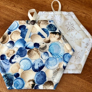 May include: Two hexagon-shaped pot holders. One is white with a beige seashell pattern and the other is white with a blue and brown seashell pattern.