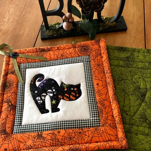 May include: A black cat with the word "Boo!" on it is the centerpiece of a Halloween-themed quilted pot holder. The pot holder is orange with a black spiderweb design and a black and white checkered border.