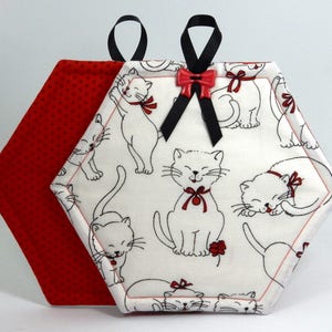 May include: White hexagon-shaped pot holder with a red polka dot backing. The front features a white background with black outlines of cats with red bows. The pot holder has black ribbon loops for hanging.