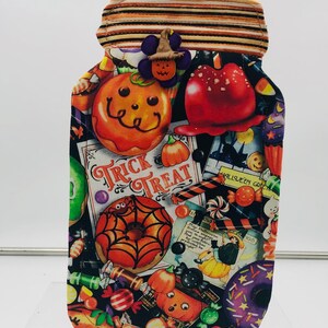 May include: A black fabric Halloween treat jar with a brown and orange striped lid. The jar is decorated with a variety of Halloween-themed images, including pumpkins, candy, donuts, and a spider web. The text "Trick Treat" is printed on the front of the jar.