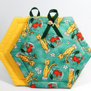 May include: A green and yellow hexagon-shaped pot holder with a cat and fish design. The pot holder has a loop on each side for hanging.