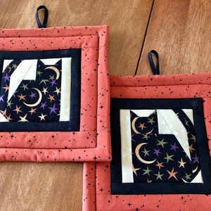 May include: Two orange and black quilted pot holders with a black cat design and gold and purple stars. The pot holders have a loop for hanging.