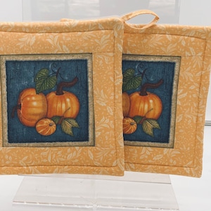 May include: Two square pot holders with a pumpkin design. The center of each pot holder features a blue square with an illustration of orange pumpkins and green leaves. The border is a light yellow fabric with a floral pattern.