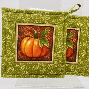 May include: Two green quilted pot holders with a brown frame and a picture of an orange pumpkin with a green stem and leaves. The pot holders have a green and white floral pattern.