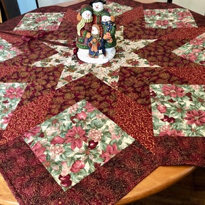 May include: A red, gold, and green quilted table runner with a star design. A snowman family figurine is in the center of the runner.