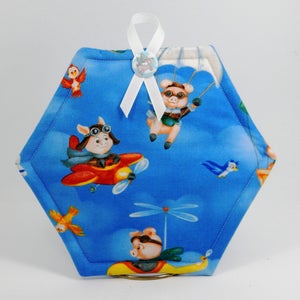 May include: Hexagon-shaped blue fabric pot holder with a white ribbon and a button featuring a pig. The fabric has a pattern of pigs flying in airplanes and hot air balloons, and birds flying in the sky.