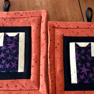 May include: Two orange and black quilted pot holders with a black cat design. The cat is made of purple fabric with white stars and is outlined in cream fabric.