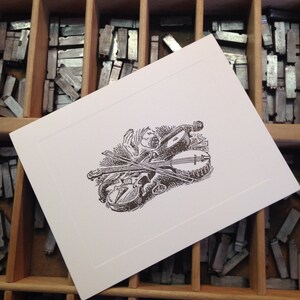 May include: A black and white illustration of a violin, banjo, and other musical instruments on a white card. The card is sitting in a wooden drawer filled with metal type.