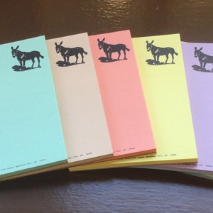 May include: A set of six rectangular paper notepads in light green, tan, peach, yellow, lavender, and cream. Each notepad features a black ink illustration of a donkey standing in the upper right corner and small printed text at the bottom.