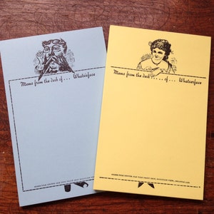 May include: Two blue and yellow memo pads with a whimsical illustration of a man and a woman on each pad. The text on the pads reads "Memo from the desk of... Whatsisface".