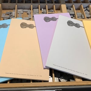 May include: Six rectangular paper notepads in pastel shades of mint green, light blue, peach, lavender, light gray, and yellow. Each notepad features a dark gray violin illustration at the top and small printed text at the bottom edge.