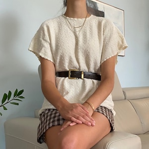 May include: A woman wearing a white crocheted sweater with a brown leather belt and a plaid skirt. She is sitting on a white couch.