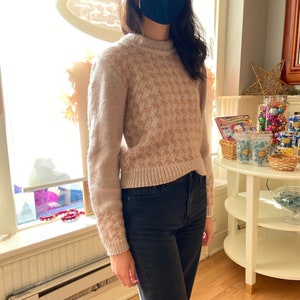 Puede incluir: Un suéter beige y marrón con estampado, cuello redondo y mangas largas. El suéter se lleva sobre unos vaqueros negros.