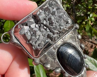 Puede incluir: Colgante de plata hecho a mano con un mineral gris texturizado grande y una piedra negra pulida. El colgante tiene un diseño geométrico único con un lazo para una cadena.