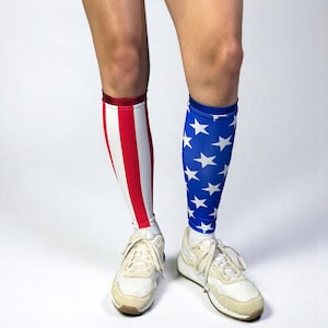 May include: A person wearing white sneakers and compression sleeves. One sleeve features red and white stripes, the other a blue background with white stars. The sleeves are worn over white socks.