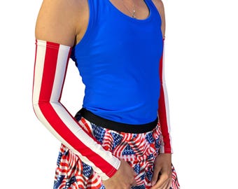 Puede incluir: Un top sin mangas azul, una falda con un estampado de la bandera estadounidense y mangas de brazo con rayas rojas y blancas. El atuendo es patriótico y adecuado para deportes o uso casual.