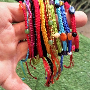 Op de afbeelding: Een stapel kleurrijke vriendschapsarmbanden met kralen. De armbanden zijn gemaakt van geweven touw en hebben verschillende kleuren, waaronder rood, geel, blauw, groen en zwart. De kralen zijn gemaakt van verschillende materialen, waaronder hout, glas en metaal.