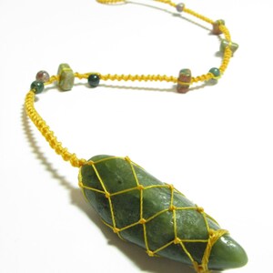 Op de afbeelding: Een groene stenen hanger met een geel koord eromheen gewikkeld in een netvormig patroon. Het koord wordt ook gebruikt om een ​​ketting te maken met verschillende andere groene, rode en zwarte stenen.