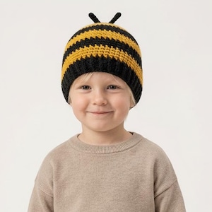 Può includere: Un bambino indossa un cappello a maglia a tema ape con strisce nere e gialle e piccole antenne. Il bambino indossa anche un maglione beige e pantaloni abbinati. L'outfit è adatto a un bambino piccolo o a un bambino piccolo.