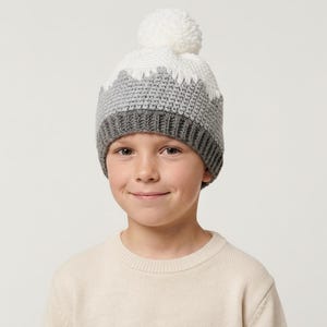 Op de afbeelding: Een gebreide wintermuts in grijstinten, wit en donkergrijs, met een witte pompon bovenop. De muts heeft een bergachtig ontwerp rond de rand. Het kind draagt een crèmekleurige trui.