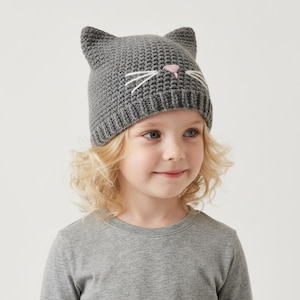 May include: A gray knit cat hat with pointed ears, white whiskers, and a pink nose. The hat is worn by a child with blonde hair and a gray long-sleeved shirt. The hat is a fun accessory.