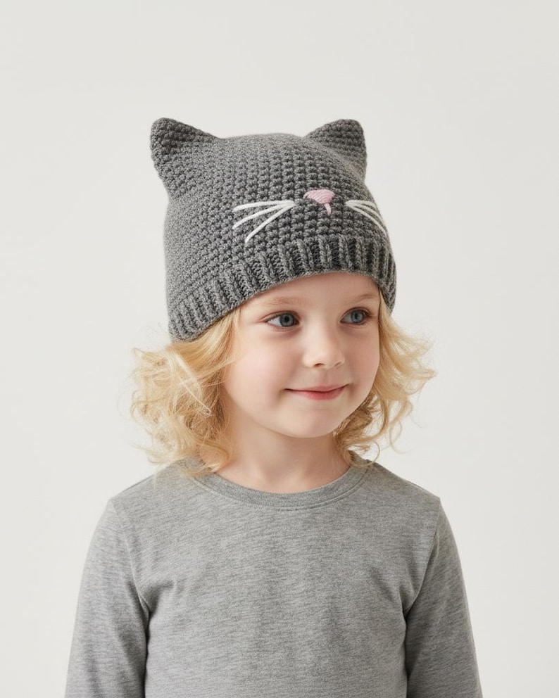 May include: A gray knit cat hat with pointed ears, white whiskers, and a pink nose. The hat is worn by a child with blonde hair and a gray long-sleeved shirt. The hat is a fun accessory.