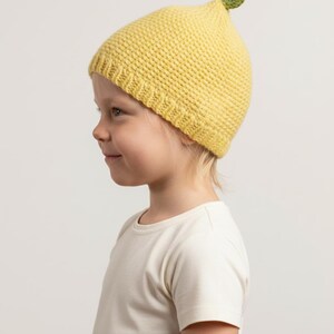 May include: A yellow knitted beanie hat featuring a textured stitch pattern and a ribbed brim. The top of the hat is adorned with a small, three-dimensional green leaf and a brown stem, designed to resemble the appearance of a lemon or fruit.