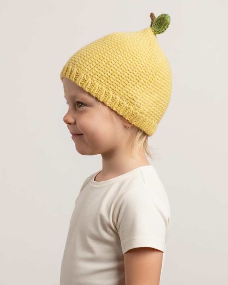 May include: A yellow knitted beanie hat featuring a textured stitch pattern and a ribbed brim. The top of the hat is adorned with a small, three-dimensional green leaf and a brown stem, designed to resemble the appearance of a lemon or fruit.