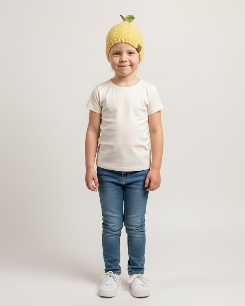 May include: A yellow knitted beanie featuring a small green leaf and brown stem detail on the crown, worn with a plain off-white short-sleeved t-shirt, blue denim jeans, and white trainers. The beanie has a ribbed texture and a small brown brand tag.