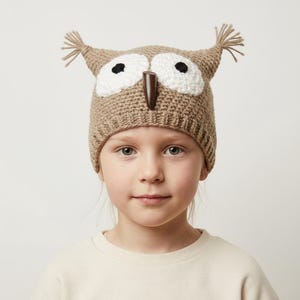 May include: A tan crocheted beanie hat featuring an owl design with two large white circular eye patches, black pupils, a dark brown vertical beak, and two small tufts of yarn at the top corners to resemble owl ears with a ribbed brim.