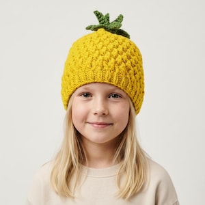 Peut inclure: Un bonnet jaune tricoté, texturé pour ressembler à un ananas, avec une couronne verte feuillue. La personne porte un pull de couleur neutre et a les cheveux blonds.