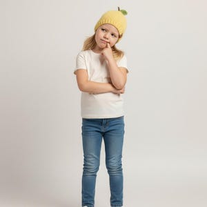 May include: A yellow knitted beanie featuring a small green leaf and brown stem detail on top, worn with a plain white short-sleeved t-shirt, blue denim jeans, and white trainers. The beanie has a ribbed texture and a rounded, fruit-like design.