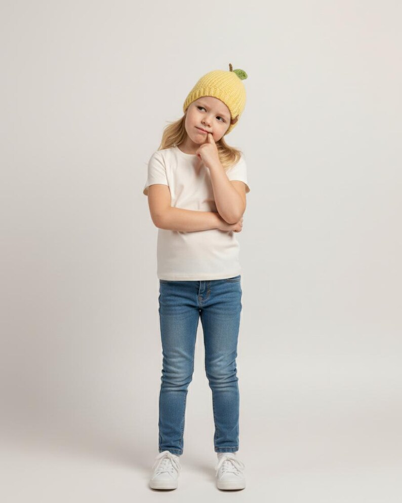 May include: A yellow knitted beanie featuring a small green leaf and brown stem detail on top, worn with a plain white short-sleeved t-shirt, blue denim jeans, and white trainers. The beanie has a ribbed texture and a rounded, fruit-like design.