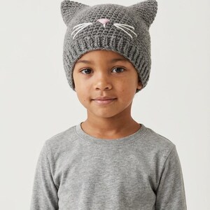 May include: A gray knit cat hat with pointed ears, white whiskers, and a pink nose. The hat is worn by a person wearing a gray long-sleeved shirt. The hat is a fun accessory for children.