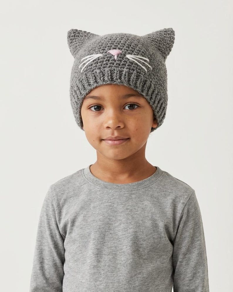 May include: A gray knit cat hat with pointed ears, white whiskers, and a pink nose. The hat is worn by a person wearing a gray long-sleeved shirt. The hat is a fun accessory for children.