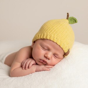 May include: A yellow crocheted hat shaped like a pear, featuring a textured knit pattern, a small brown stem, and a single green leaf on top. The hat includes a ribbed brim and a small brown leather-like tag attached to the side edge.