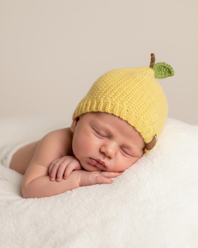 May include: A yellow crocheted hat shaped like a pear, featuring a textured knit pattern, a small brown stem, and a single green leaf on top. The hat includes a ribbed brim and a small brown leather-like tag attached to the side edge.